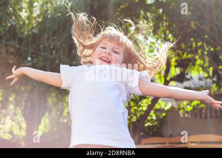 Glückliches kleines Mädchen springen aufgeregt, fliegende Haare, Arme weit verbreitet Nahaufnahme. Fröhlich enthusiastisch lachendes Kind springen im Sonnenlicht, Glück, Spaß Stockfoto