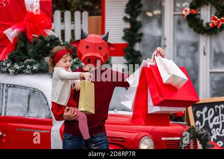 Ein junger Mann in einer roten Stiermaske hält ein kleines Baby, weiße Pakete. Neujahrsbummel, Verkauf. Die Familie steht am Auto, Bäume im Schnee Stockfoto