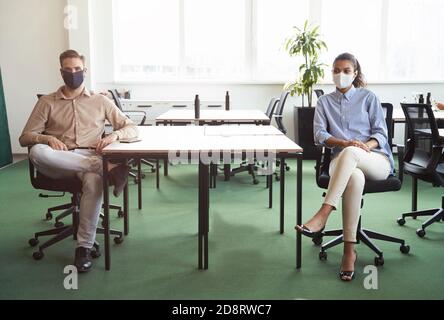Zwei junge, vielfältige Kollegen tragen eine schützende Gesichtsmaske beim Betragen Kamera, während Sie am Schreibtisch im modernen Büro sitzen Stockfoto
