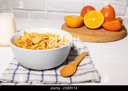 Maisflocken Schüssel mit Milch, Persimmon, Orange und Holzlöffel auf weißem Hintergrund, gesundes Frühstück Stockfoto