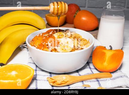 Maisflocken Schüssel mit Banane, Milch, Persimmon, Orange und Honig auf weißem Hintergrund, gesundes Frühstück Stockfoto