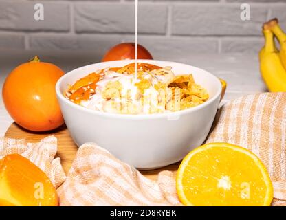 Maisflocken Schüssel mit Milch, Persimmon, Orange und Holzlöffel auf weißem Hintergrund, gesundes Frühstück Stockfoto