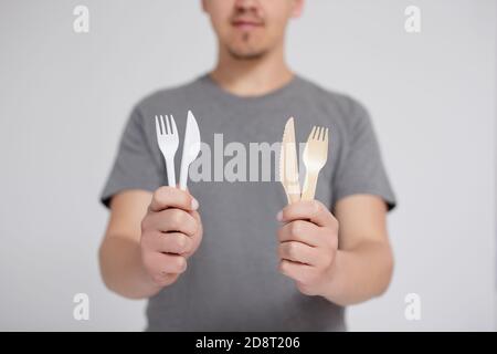 Zero Waste und umweltfreundliches Konzept - junger Mann im Vergleich Gabeln und Messer aus Kunststoff und Holz auf grauem Hintergrund Stockfoto