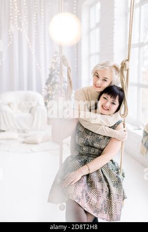 Schöne kaukasische Mädchen in Kleid, sitzt auf der Schaukel in gemütlichen Licht dekoriert Studio mit Weihnachten und Neujahr Dekorationen, Tannenbaum und großes Fenster Stockfoto