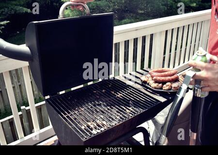 Offener Holzkohlegrill auf einem Balkon, mit einem Teller gekochter Wurst und Huhn auf der Seite. Mit einer Person, die Zangen hält und sich bereit, den Deckel zu schließen. Stockfoto