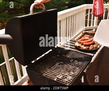 Offener Holzkohlegrill auf einem Balkon, mit einem Teller gekochter Wurst und Huhn auf der Seite. Mit einer Person, die Zangen hält und sich bereit, den Deckel zu schließen. Stockfoto