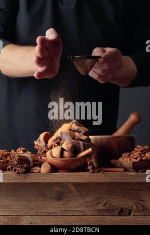 Der Koch streut die Schokolade mit Kakaopulver. Auf einem alten Holztisch Süßigkeiten, Kaffeebohnen, Zimt, Anis, Nüsse und Stücke von gebrochenem schwarzem Choco Stockfoto