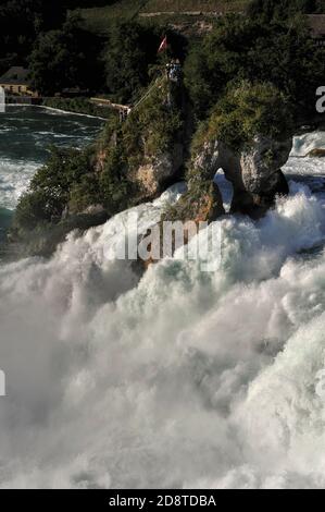 Die Schweizer Flagge fliegt vom Aussichtspunkt auf den Rheinfallfelsen, einem felsigen Felsen, der auf beiden Seiten vom donnernden Rheinfall, dem Rheinfall bei Schaffhausen, der Nordschweiz, dem größten und mächtigsten Wasserfall Europas, passiert wird. Der Rheinfallfelsen, der nur mit dem Boot erreicht wurde, war Teil einer harten Kalksteinklippe, die den ehemaligen Kanal des hoch- oder Oberrheins flankiert. Trotz Erosion hat es Sommerwasserströmungen mit durchschnittlich 600 Kubikmeter (21,000 Kubikfuß) pro Sekunde standgehalten. Der Fluss stürzt etwa 23 m (75 ft), bevor er sich sediger in Richtung Deutschland entwickelt. Stockfoto