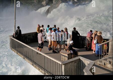 Eine Aussichtsplattform, die vom Ufer unterhalb von Schloss Laufen ragt, bietet den Touristen einen atemberaubenden Blick auf Europas größten und mächtigsten Wasserfall, den Rheinfall oder Rheinfall, bei Schaffhausen, Nordschweiz. Der hoch- oder Oberrhein donnert hier mit durchschnittlich 600 Kubikmetern (21,000 Kubikfuß) pro Sekunde über ein felsiges Flussbett. Die spektakulären Wasserfälle sind eine beliebte Touristenattraktion und ziehen jedes Jahr Zehntausende von Touristen an. Stockfoto