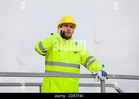 Junger Ingenieur oder Arbeiter, der vor dem Telefon spricht Von Windenergieanlagen im unscharfen Hintergrund Stockfoto