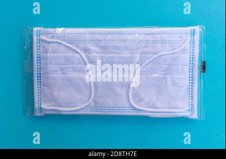 Flach legen einer Einweg-Covid-19 Coronavirus schützende chirurgische Gesichtsmaske, verpackt in einer Plastiktüte. Auf türkisblauem Hintergrund Stockfoto