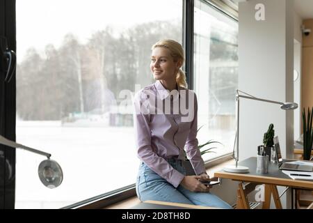 Verträumte junge Geschäftsfrau sitzt auf Fensterbank im Büro. Stockfoto