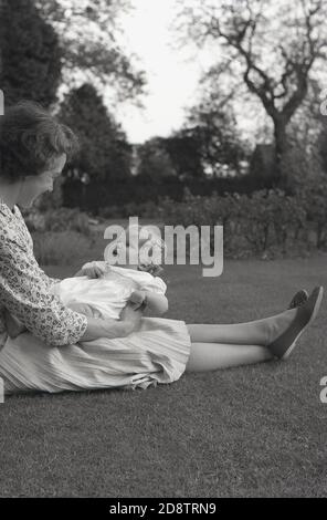 1950s, historisch, draußen auf einem Rasen, eine Mutter, die auf dem Gras sitzt und ihr Baby auf ihrem Schoß hält und sie lächelnd ansieht, England, Großbritannien. Stockfoto