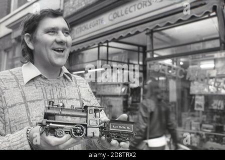 1980s, historisch, vor einem Modelleisenbahn- und Sammlergeschäft in der Micklegate Street, York, England, Großbritannien, ein Mann, der stolz ein Modell einer L & N W R Dampflokomotive und eines Wagens hält. Modelleisenbahnen sind ein beliebtes Hobby und diejenigen, die Modelleisenbahnen sammeln, sind als Eisenbahnenthusiasten bekannt. In der englischen Stadt York befindet sich das National Railway Museum, in dem sich weltverändernde Erfindungen wie die Rocket und die Mallard, die schnellste Dampflokomotive der Welt, befinden. Stockfoto