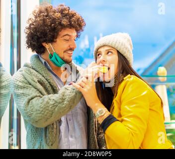 Junges und glückliches Paar trägt schützende Gesichtsmaske essen ein Makkaronen stehen in einem Shop.Girl mit einem süßen Dessert Süßigkeiten Im Herbst.Neu normal Stockfoto