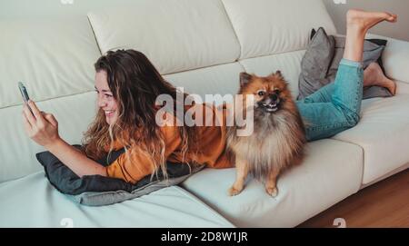 Lächelnde Frau mit dem Telefon, das mit ihrem Hund auf der Couch liegt. Stockfoto