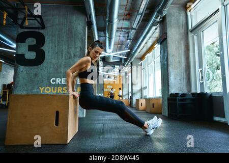 Fitnesstraining. In voller Länge junge athletische Frau in Sportbekleidung tun Trizeps Übungen auf hölzernen Crossfit Jump Box im Fitnessstudio. Sportliche Menschen, gesunder Lebensstil und Workout-Konzept Stockfoto