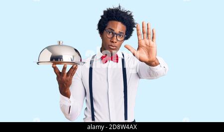 Hübscher afroamerikanischer Mann mit afro-Haaren in Kellneruniform Halten silberne Tablett mit offener Hand tun Stop-Zeichen mit Seriöse und selbstbewusste expr Stockfoto