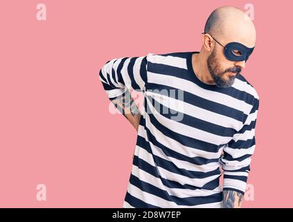 Junger, schöner Mann mit Einbrechermaske, der Rückenschmerzen hat, den Rücken mit der Hand berührt, muskuläre Schmerzen Stockfoto