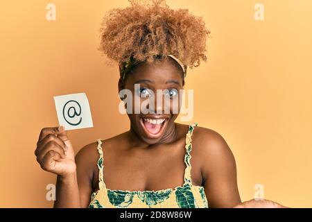 Junge afrikanische Frau mit afro Haar halten Online-Mail-Symbol Auf dem Papier feiert Leistung mit glücklichen Lächeln und Gewinner Ausdruck Mit erhobener Hand Stockfoto