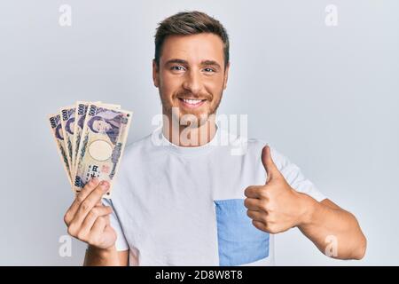 Schöner kaukasischer Mann hält 5000 japanische Yen-Banknoten lächeln glücklich und positiv, Daumen nach oben tun ausgezeichnet und Genehmigung Zeichen Stockfoto