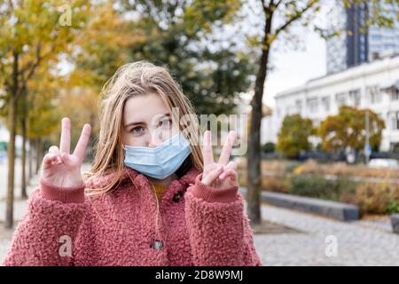 Mädchen trägt Gesichtsmaske für Corona-Virus mit positiver Einstellung. Friedenszeichen mit zwei Händen geben. Stockfoto