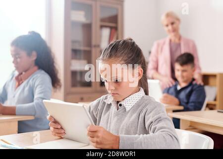 Nettes Mädchen und ihre Klassenkameraden mit Tabletten arbeiten individuell mit Online-Text Stockfoto