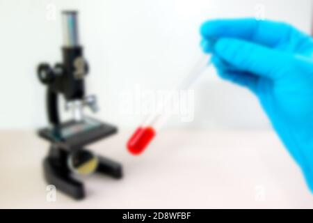 Unschärfe eines Mikroskops und einer blauen Hand mit Handschuhen, die ein Reagenzglas mit roter Flüssigkeit hält. Das Reagenzglas wird in einem Winkel gehalten. Mikroskop ist im Hintergrund. Stockfoto