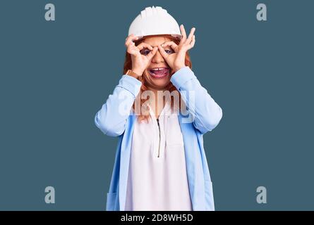 Junge lateinische Frau trägt Architekt Hardhut tun ok Geste wie Fernglas herausstreckt Zunge, Augen durch die Finger schauen. Verrückter Ausdruck. Stockfoto