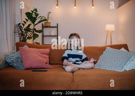 Ruhiges kleines Mädchen, das auf einer Couch in voller Lotusposition sitzt. Meditieren Stockfoto