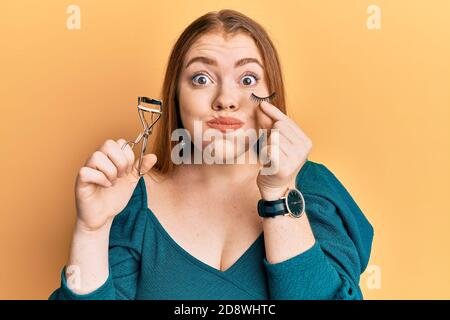 Junge schöne Rothaarige Frau hält Wimpern Lockenwickler und gefälschte Wimpern Puffing Wangen mit lustigen Gesicht. Mund mit Luft aufgeblasen, Luft fangen. Stockfoto