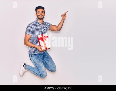 Junge gutaussehende hispanische Mann trägt Freizeitkleidung lächelnd glücklich. Springen mit Lächeln auf Gesicht hält Geburtstagsgeschenk mit Finger auf die Seite OV zeigen Stockfoto