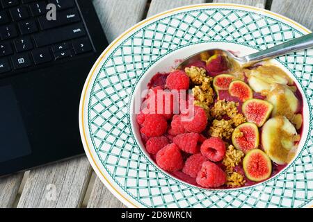 Essen Acai-Schale mit frischen Feigen, Himbeeren, Bananen und Müsli während der Arbeit an einem Laptop-Computer Stockfoto