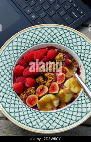 Essen Acai-Schale mit frischen Feigen, Himbeeren, Bananen und Müsli während der Arbeit an einem Laptop-Computer Stockfoto