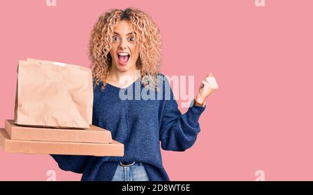 Junge blonde Frau mit lockigen Haaren hält Lieferung Essen schreien stolz, feiert Sieg und Erfolg sehr aufgeregt mit erhobenen Armen Stockfoto