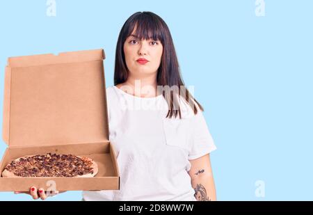 Junge plus size Frau hält Lieferung Pizza Box Denken Haltung Und nüchterner Ausdruck, der selbstbewusst aussieht Stockfoto