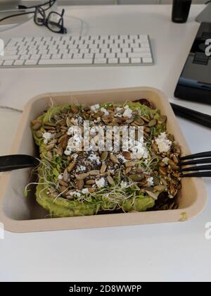 Mittagessen in einem Büro. Zertrümmerte Avocado-Toast mit Feta, Sonnenblumenkernen und Luzerne. Sandwich auf einem Computertisch mit Tastatur Stockfoto