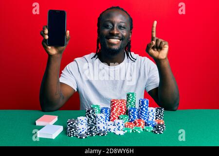 Hübscher junger schwarzer Mann spielt Poker hält Smartphone lächelnd mit einer Idee oder Frage Zeigefinger mit glücklichem Gesicht, Nummer eins Stockfoto