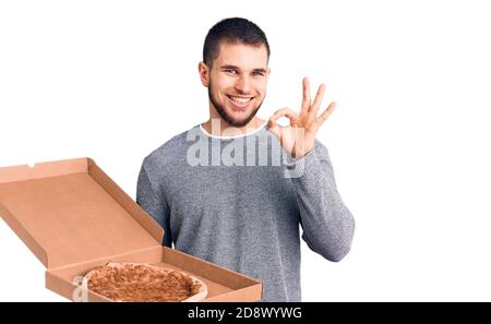 Junge gut aussehende Mann hält Lieferung italienische Pizza tun ok Zeichen mit Fingern, lächelnd freundlich gestikulieren ausgezeichnete Symbol Stockfoto