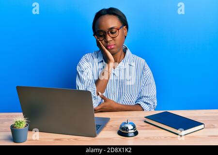 Junge afrikanerin, die an der Hotelrezeption mit einem Laptop arbeitet und dabei denkt, dass sie müde und gelangweilt aussieht mit Depressionen, die mit gekreuzten Armen zu kämpfen haben. Stockfoto