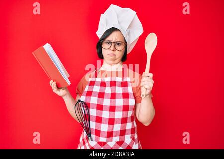 Brunette Frau mit Down-Syndrom tragen professionelle Bäcker Schürze lesen Kochen Rezept Buch ahnungslos und verwirrt Ausdruck. Zweifel Konzept. Stockfoto