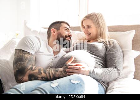 Der Mann lehnte sich an den Babybauch seiner schwangeren Frau, die auf dem Bett liegt Stockfoto