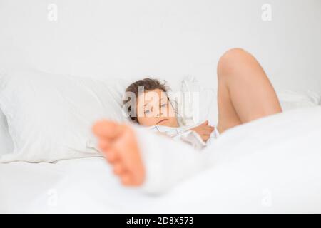 Frakturen der Beine des Kindes. Ein Teenager-Mädchen ruht nach der ersten Hilfe auf der orthopädischen Station auf der Station. Ein Baby Bein in Gips auf einem weißen Bett Stockfoto