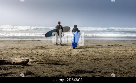 Zwei Surfer auf dem Weg in den mit Nebel bedeckten Pazifik am Cox Bay Beach im Pacific Rim National Park auf Vancouver Island, British Columbia, Kanada Stockfoto