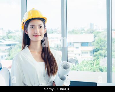Asiatische glücklich Business Frau Ingenieur Entwickler tragen gelben Schutzhelm mit Blaupause Papiere auf Fenster Glas Hintergrund mit Kopierer Raum. Archit Stockfoto
