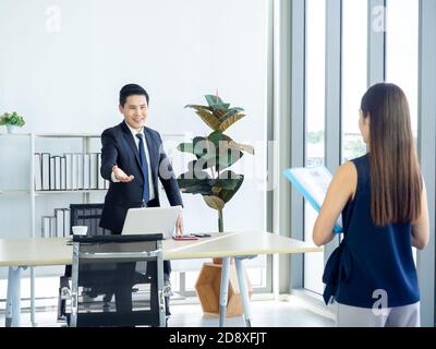 Asiatischer Geschäftsmann, Chef in Anzug macht Hand Geste, um junge Frau halten Lebenslauf in der Job-Interview einladen, um sich auf dem Stuhl in der Nähe von riesigen Glas Stockfoto