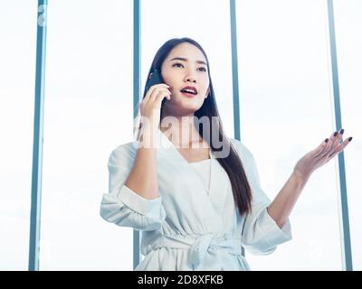 Junge hübsche asiatische Frau trägt formell weißes Hemd im Gespräch auf dem Telefonanruf in der Nähe von riesigen Glasfenster im Büro. Porträt der schönen Geschäftsfrau U Stockfoto