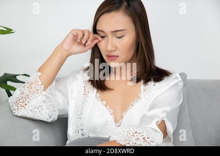 Asiatische Frauen in einem Nachtkleid und tragen weiße Satin-Robe, die nachts auf dem grauen Sofa im Wohnzimmer sitzt, EIN Mädchen in SeidenNachtwäsche sc Stockfoto