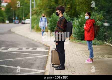 Junge Leute mit Taschen warten auf Bushaltestelle im Freien in der Stadt. Coronavirus-Konzept. Stockfoto