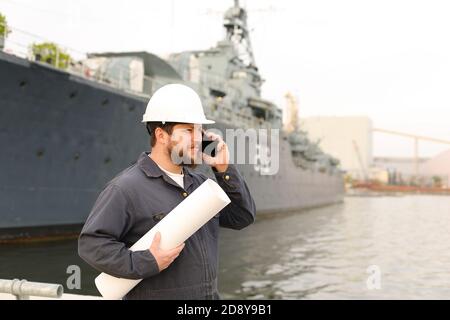 Marine männliche Deckhand im Gespräch mit VHF Walkie Talkie, hält Papiere in der Nähe des Schiffes im Hintergrund. Stockfoto